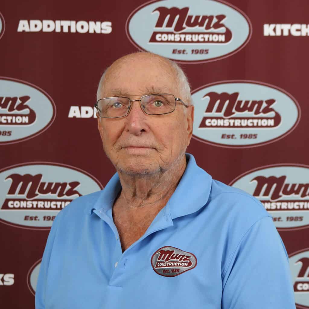 An older man wearing glasses and a light blue Munz Construction polo shirt stands in front of a maroon Munz Construction backdrop featuring the company logo and words like Additions and Kitchens.