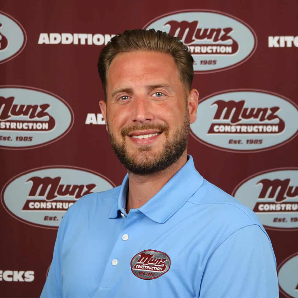 A man with short brown hair and a beard, wearing a light blue Munz Construction polo shirt, smiles in front of a maroon Munz Construction backdrop with white text and logos.