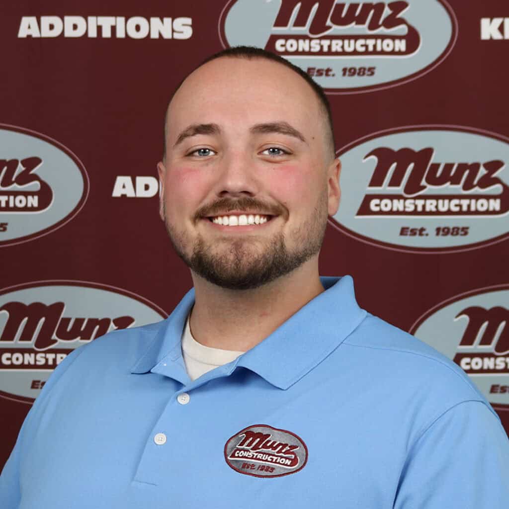 A smiling man with a short beard wearing a light blue Munz Construction polo shirt stands in front of a maroon Munz Construction backdrop.