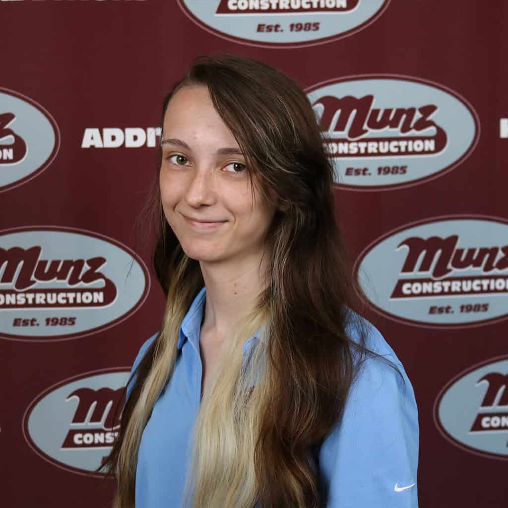 A young woman with long brown and blonde hair wearing a light blue collared shirt stands in front of a maroon Munz Construction banner. She is smiling slightly and facing the camera.