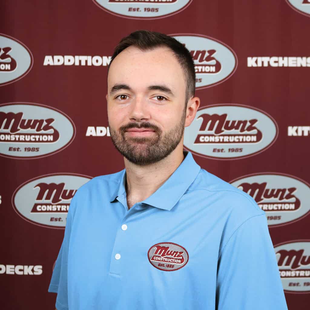 A man with short dark hair and a beard wears a light blue polo shirt with a Munz Construction logo. He stands in front of a maroon backdrop displaying the Munz Construction logo and various service words.