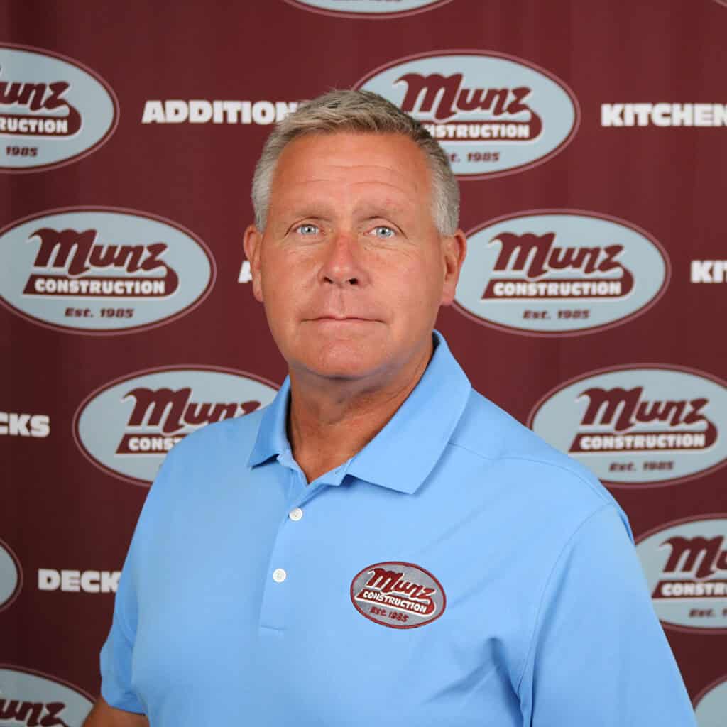 A man in a light blue polo shirt with a Munz Construction logo stands in front of a maroon backdrop featuring repeated Munz Construction logos and words like ADDITIONS, KITCHENS, and DECKS.
