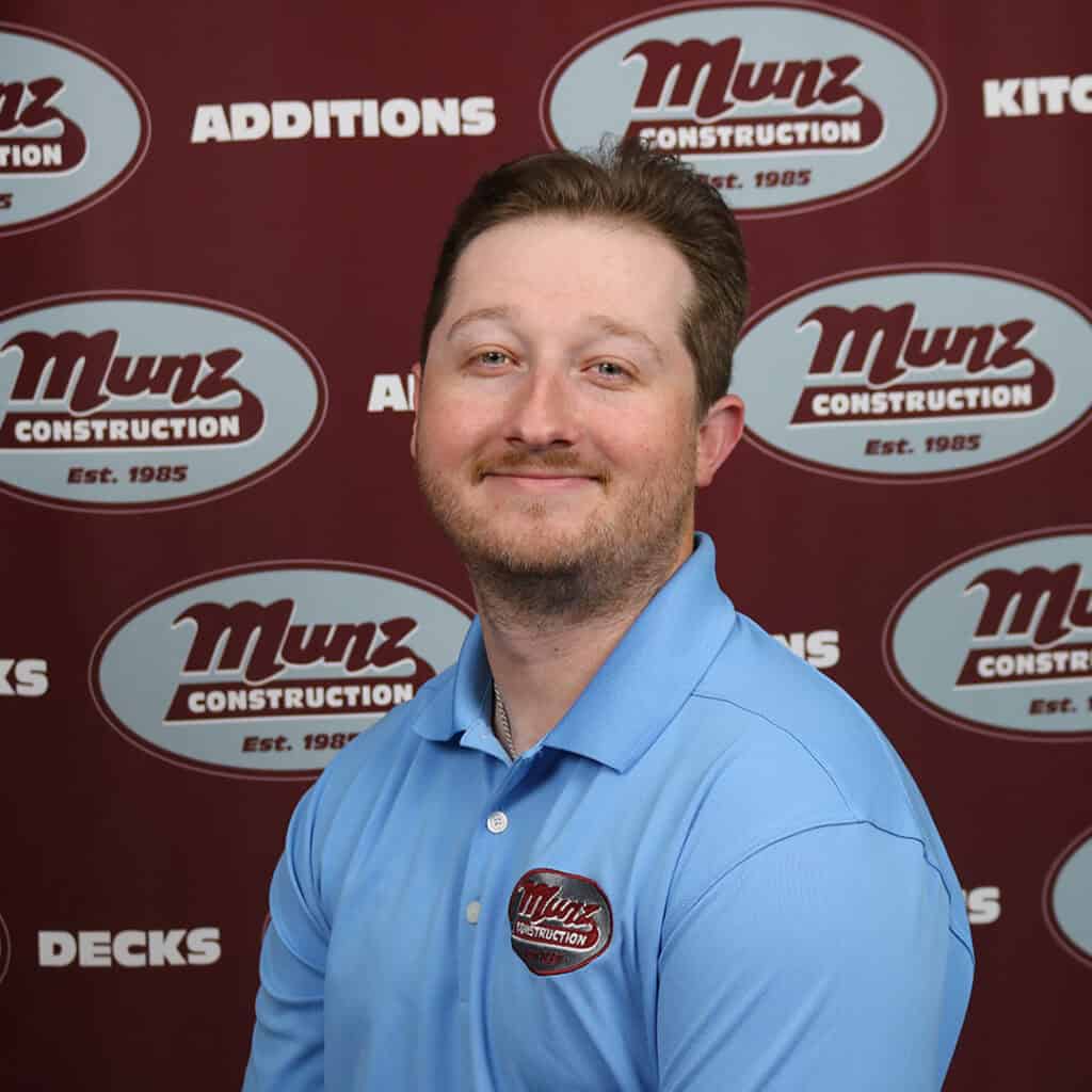 A man with short hair and a beard, wearing a light blue Munz Construction polo, smiles in front of a maroon backdrop with repeated Munz Construction logos and words like ADDITIONS and DECKS.