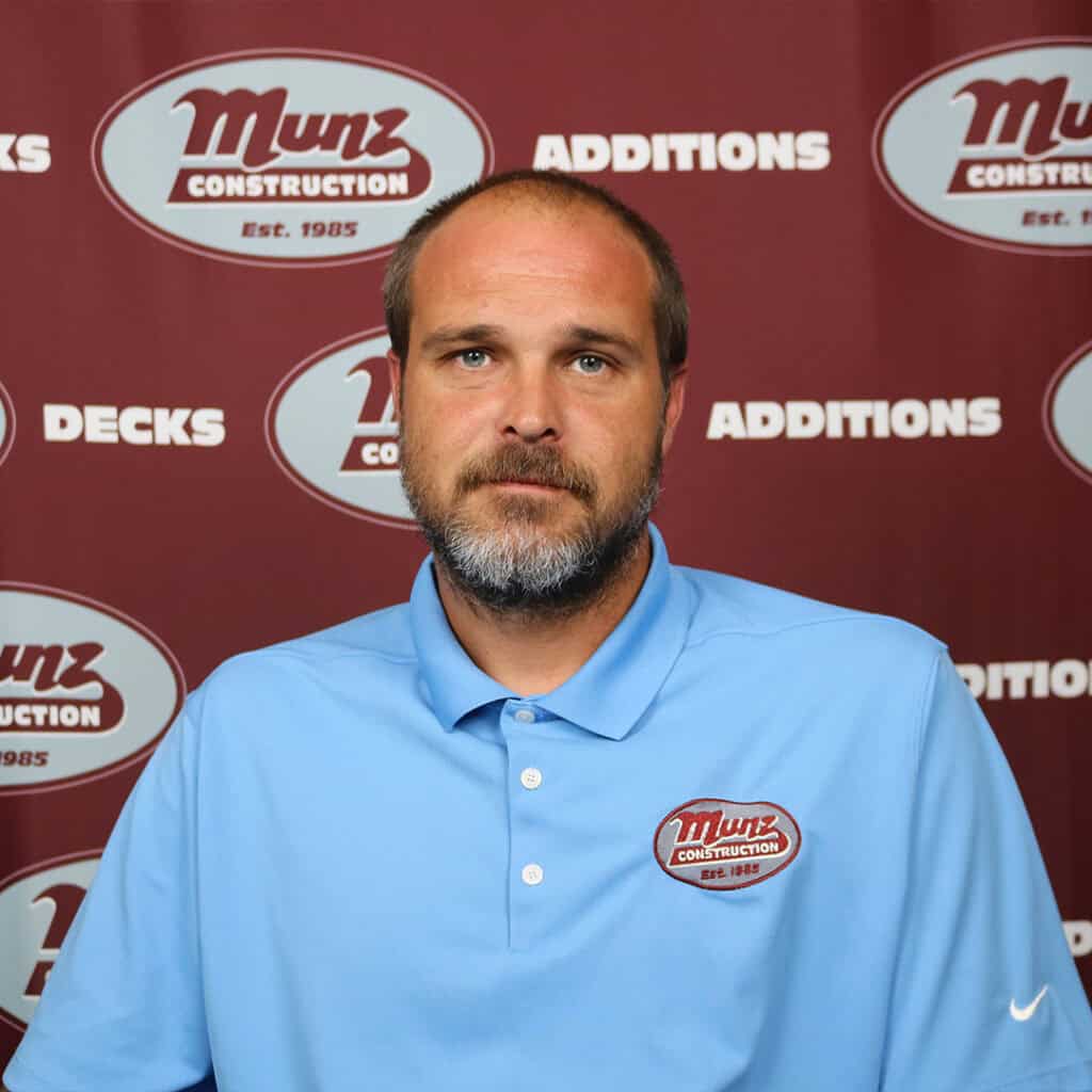 A man with a beard wearing a light blue Munz Construction polo shirt sits in front of a red Munz Construction backdrop with logos and words like Additions and Decks.