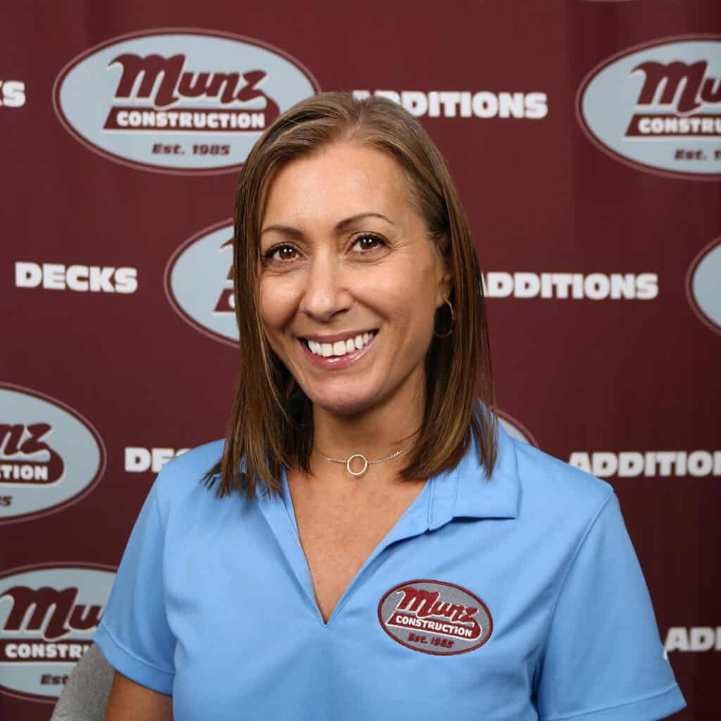 A woman with straight brown hair wearing a light blue Munz Construction polo shirt smiles in front of a maroon Munz Construction backdrop.