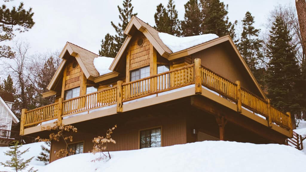A wooden cabin with a wraparound balcony sits on a snow-covered hillside, surrounded by tall evergreen trees under an overcast sky. Stay warm with these winter home upgrades!.