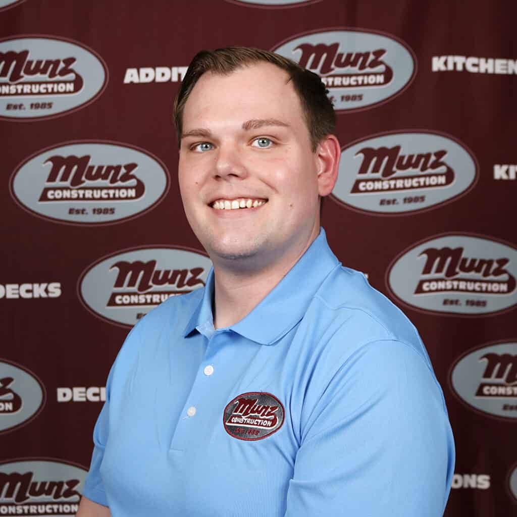 A smiling man in a light blue Munz Construction polo shirt poses in front of a maroon Munz Construction-branded backdrop.