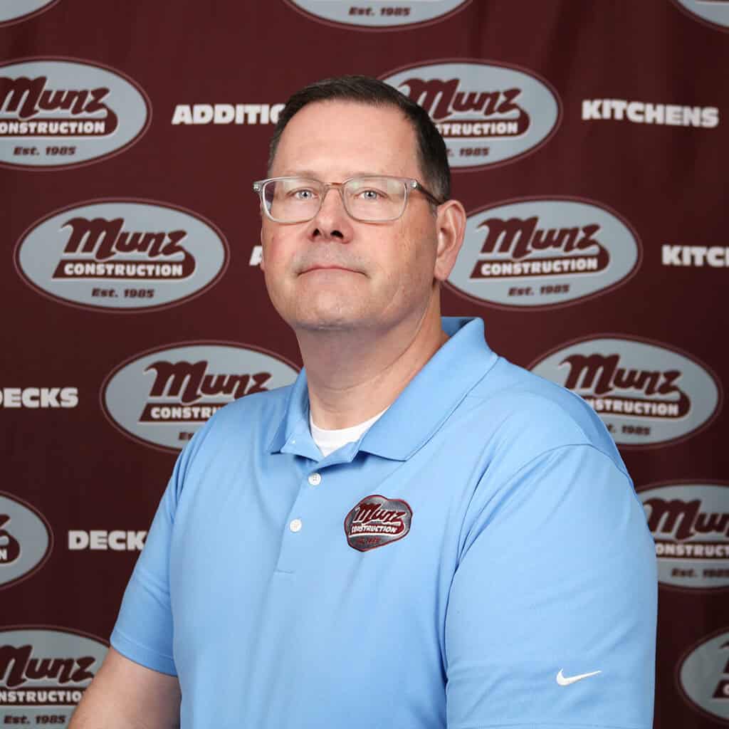 A man wearing glasses and a light blue Munz Construction polo shirt poses in front of a maroon Munz Construction step-and-repeat backdrop.