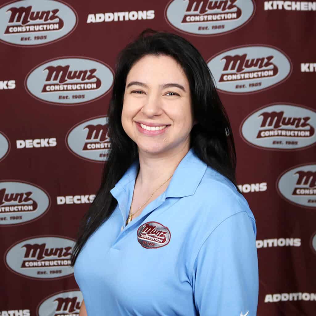 A woman with long dark hair smiles while wearing a light blue Munz Construction polo shirt, standing in front of a maroon Munz Construction backdrop with words like Decks, Additions, and Kitchens.