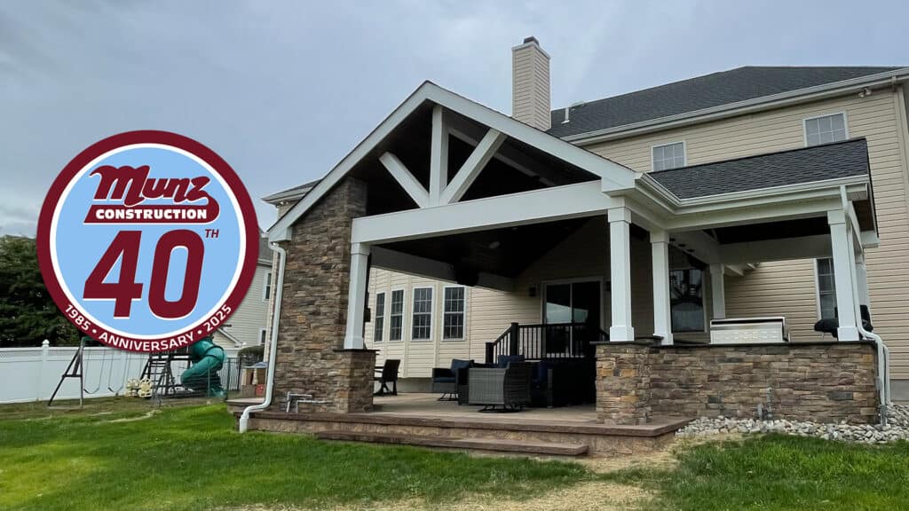 Get Summer-Ready with this beige house featuring a large covered patio and outdoor seating. The red and white Munz Construction 40th Anniversary 1985-2025” logo stands out in the left foreground, and a playset sits in the fenced yard.