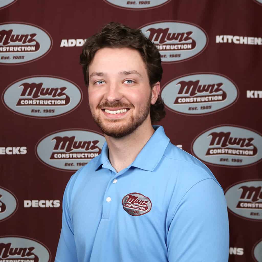 A smiling man with brown hair and a beard wearing a light blue polo shirt with a Munz Construction logo, standing in front of a Munz Construction-branded backdrop.