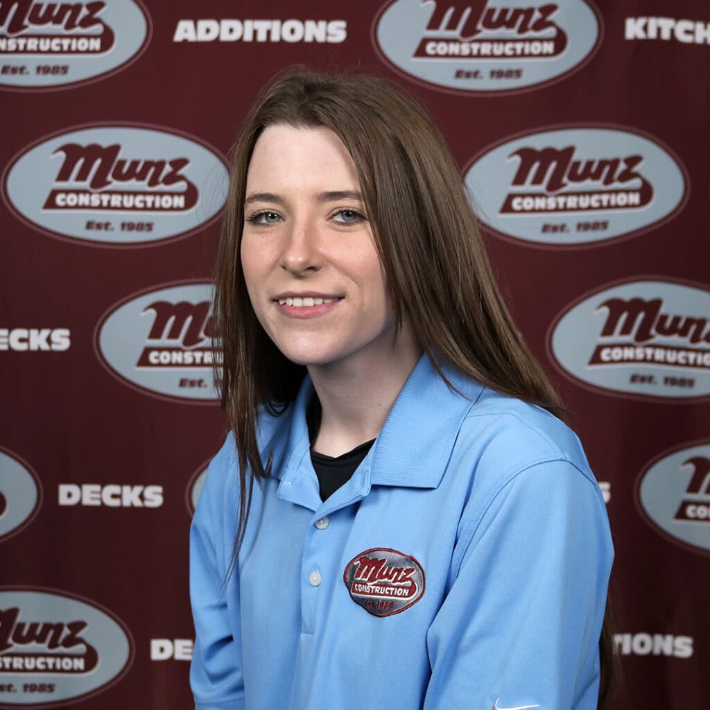 A woman with long brown hair wearing a light blue Munz Construction polo shirt smiles at the camera in front of a Munz Construction branded backdrop.