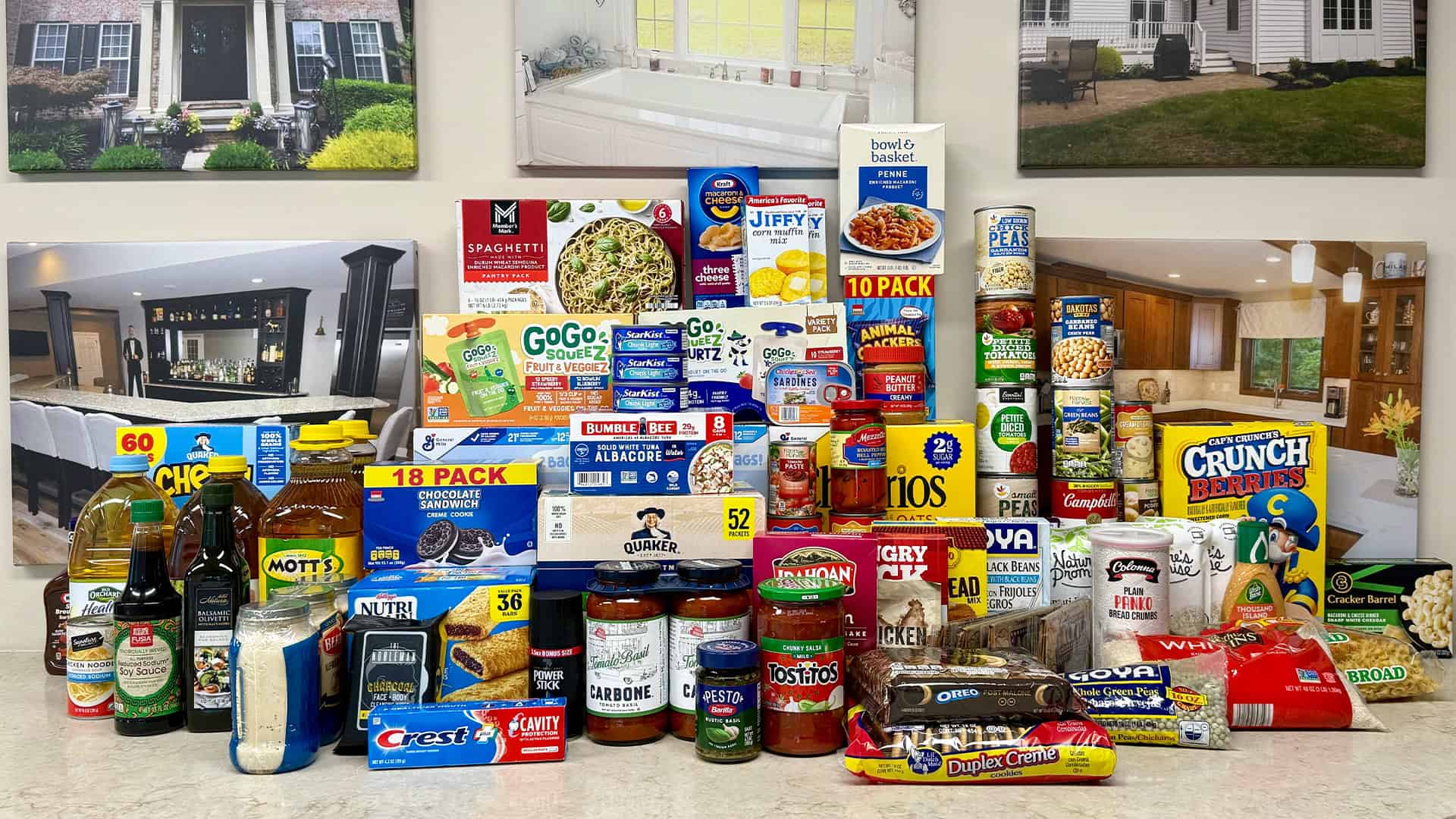 A large assortment of non-perishable food items and household essentials collected for a food drive, displayed on a countertop with home interior photos in the background.
