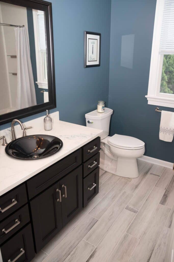 A modern bathroom with blue walls, a white toilet, dark wood vanity with a black glass sink, white marble countertop, towel ring, framed art, and wood-look tile flooring. A window lets in natural light.