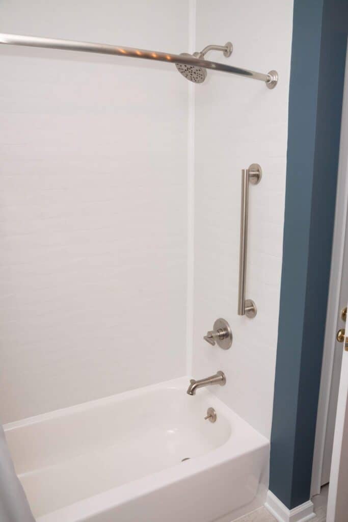 A clean white bathtub with a showerhead, a curved shower curtain rod, a silver grab bar, and silver fixtures against white walls and a blue accent wall near the bathroom door.