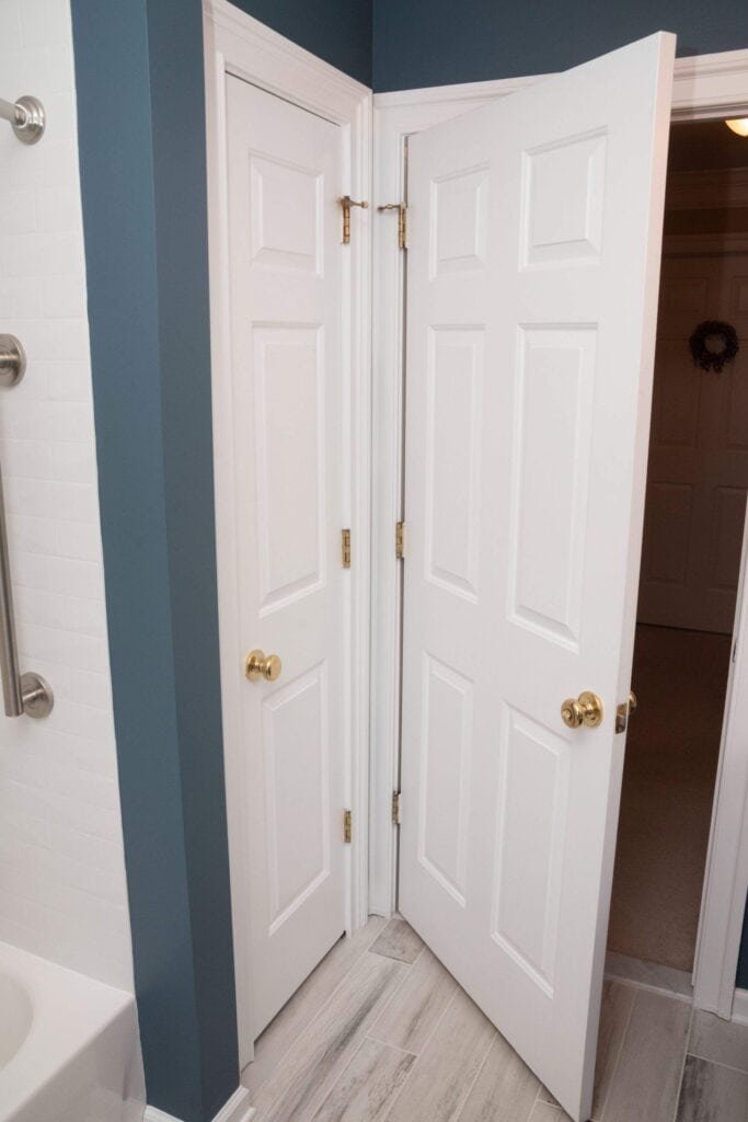 Two white doors are installed at a 90-degree angle in a small corner, with both doors open and nearly touching each other. The walls are blue, and the floor is light-colored tile. A bathtub and grab bar are visible on the left.