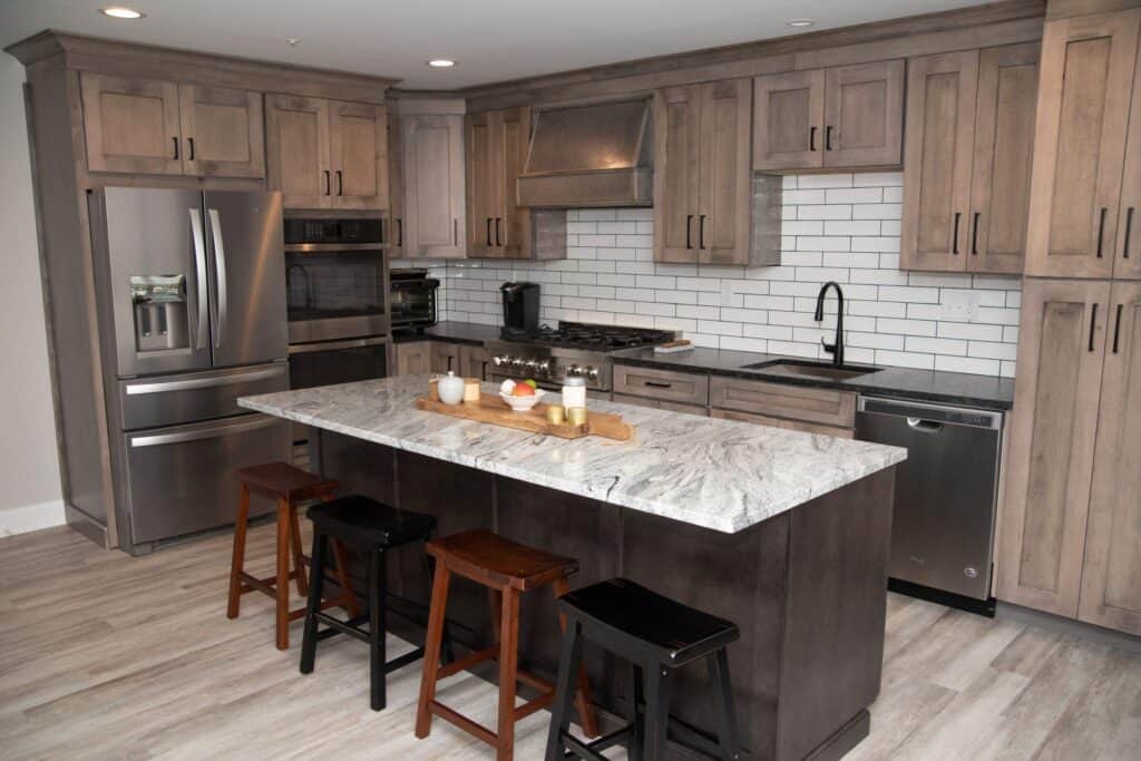 Modern kitchen with wood cabinets, stainless steel appliances, white subway tile backsplash, and a marble island with three stools. The island has a wooden tray holding cups and a bowl. The floor is light-colored wood.