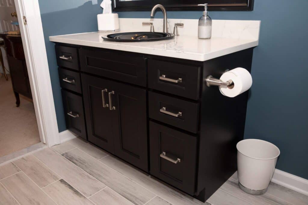 A modern bathroom vanity with black cabinets, silver handles, a white countertop, a tissue box, a soap dispenser, a sink, a mounted toilet paper roll, and a white trash can on gray wood-look tile flooring.