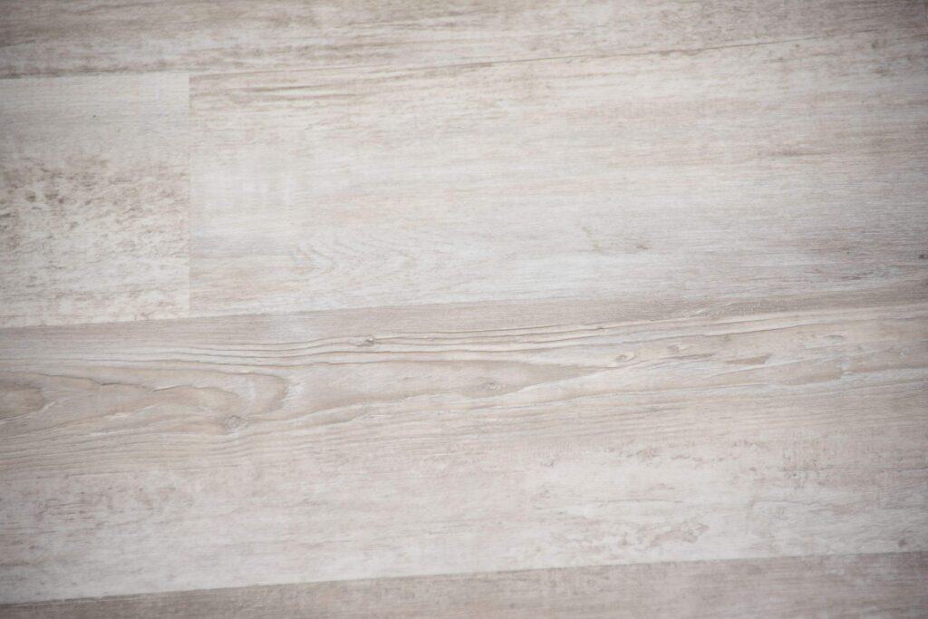 Light gray wooden laminate flooring with subtle grain patterns and a smooth, matte finish, showing variations in tone and natural wood texture.