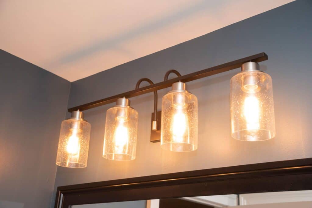 A modern wall-mounted light fixture above a mirror features four illuminated bulbs, each enclosed in a clear cylindrical glass shade, set against a dark gray wall.