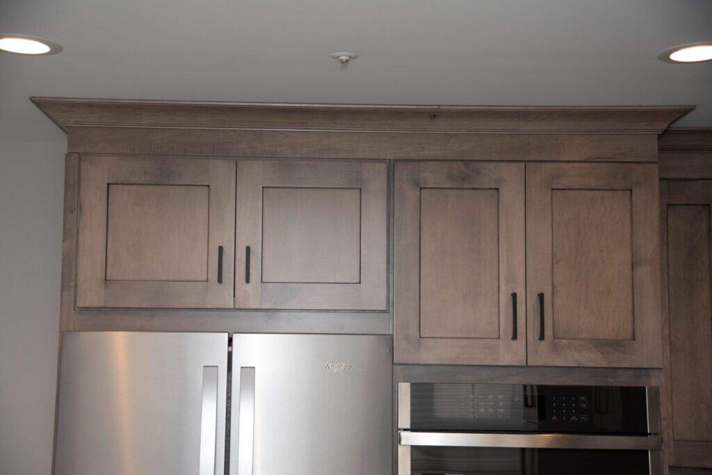 Upper kitchen cabinets in a light wood finish with black handles are above a stainless steel refrigerator and built-in microwave, set against a plain light-colored wall and ceiling with recessed lighting.