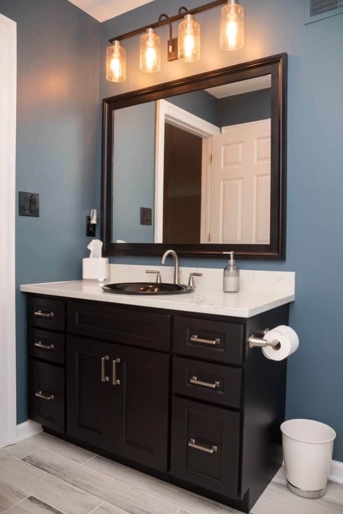Modern bathroom with dark wood vanity, white countertop, large mirror, trio of glass light fixtures, soap dispenser, tissue box, toilet paper holder, and white trash bin against blue walls and light gray tile floor.