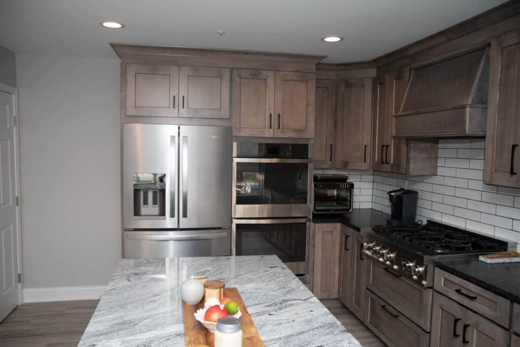 Modern kitchen with gray wood cabinets, stainless steel refrigerator, double wall oven, gas stove, white subway tile backsplash, and a marble island countertop with a wooden tray holding fruit and condiments.