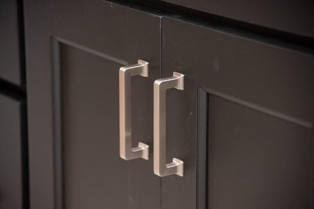 Close-up of two metallic handles on dark-colored cabinet doors, showing the geometric design and smooth finish of the hardware against the paneled surface.