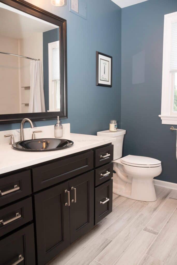 Modern bathroom with blue walls, white trim, a dark wood vanity with a black sink, silver fixtures, a white toilet, light wood-look flooring, a wall mirror, and a framed picture above the toilet.