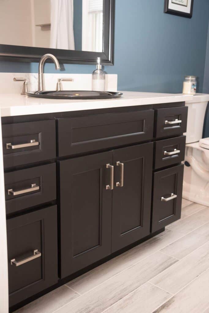 Modern bathroom with dark wood vanity, silver handles, a sink with a silver faucet, soap dispenser, and a mirror above. Blue walls, light wood-look tile flooring, and part of a toilet are visible.