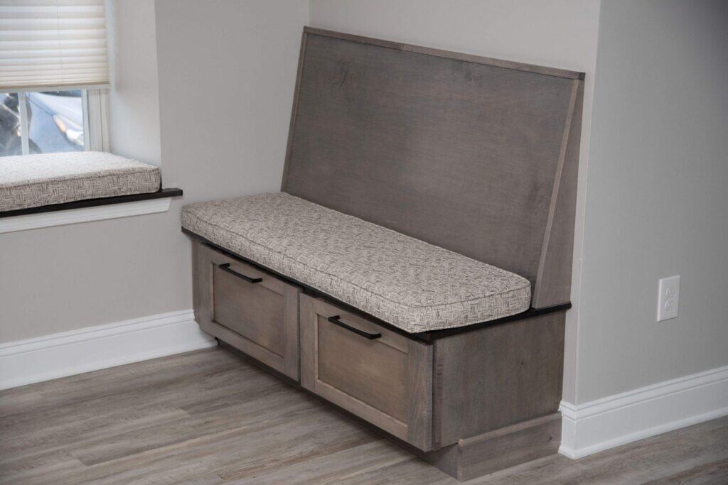 A gray wooden bench with a patterned cushion sits against a wall; the bench has two drawers with black handles and is next to a window with a matching cushioned seat.