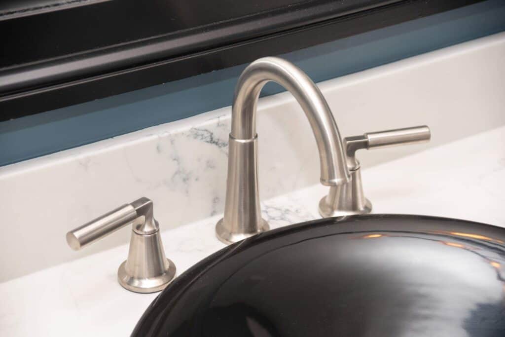 A close-up of a modern bathroom sink with a brushed nickel faucet and two handles on a white marble countertop, next to a black sink basin.