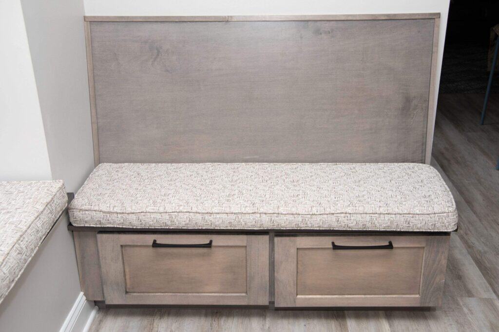 A wooden bench with a cushioned seat and two drawer handles beneath, placed against a wall on a light-colored wood floor.