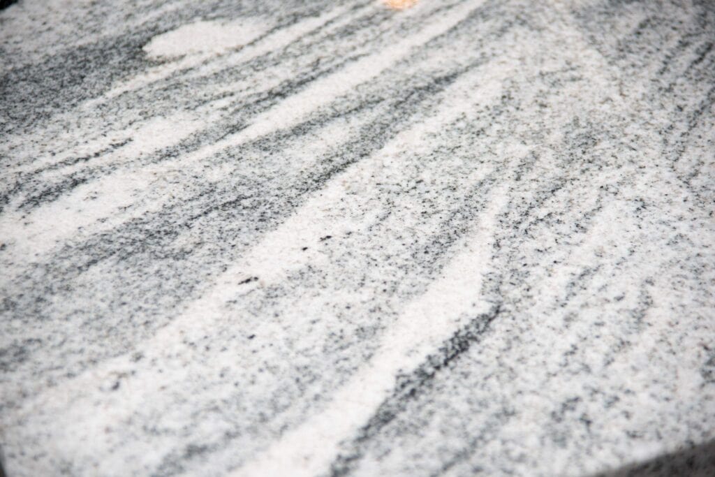 Close-up of a polished granite surface with gray, white, and black streaks forming a natural, swirling pattern. The texture appears smooth and slightly reflective.