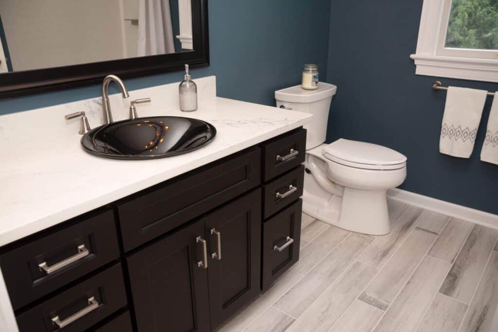 A modern bathroom with a dark vanity, black vessel sink, silver faucet, white toilet, patterned towels, and light gray wood-look tile flooring against blue walls. A candle sits on the toilet tank.