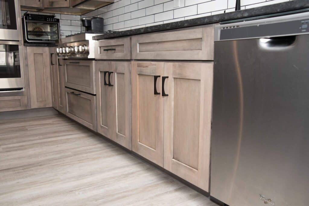 Modern kitchen with light wood cabinets, black handles, stainless steel dishwasher, and stove. The backsplash features white subway tiles, and the floor has a light wood finish. Countertops are dark and sleek.