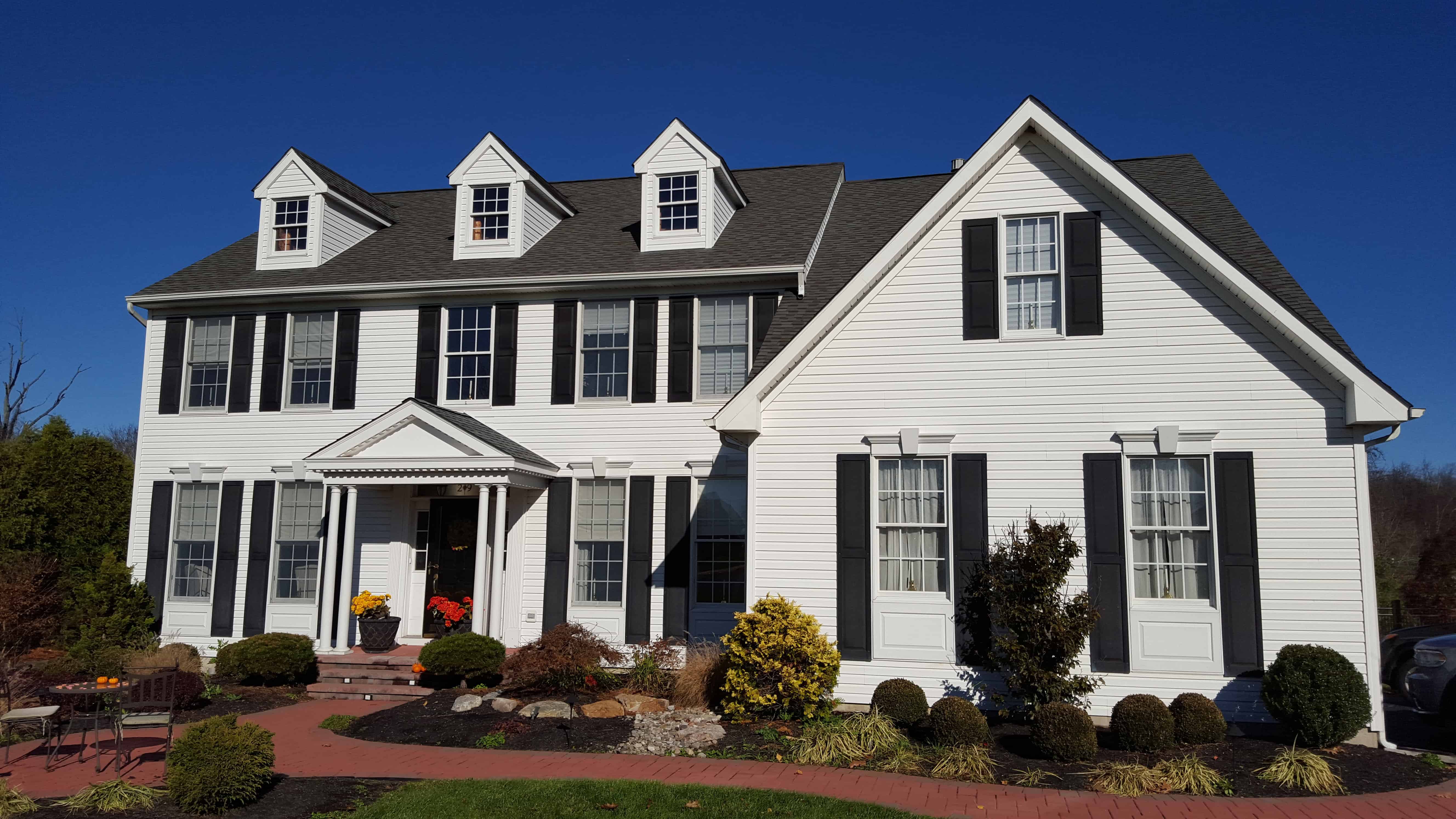 A large, two-story white house with black shutters, three dormer windows, a covered front entrance, and neatly landscaped garden beds under a clear blue sky.