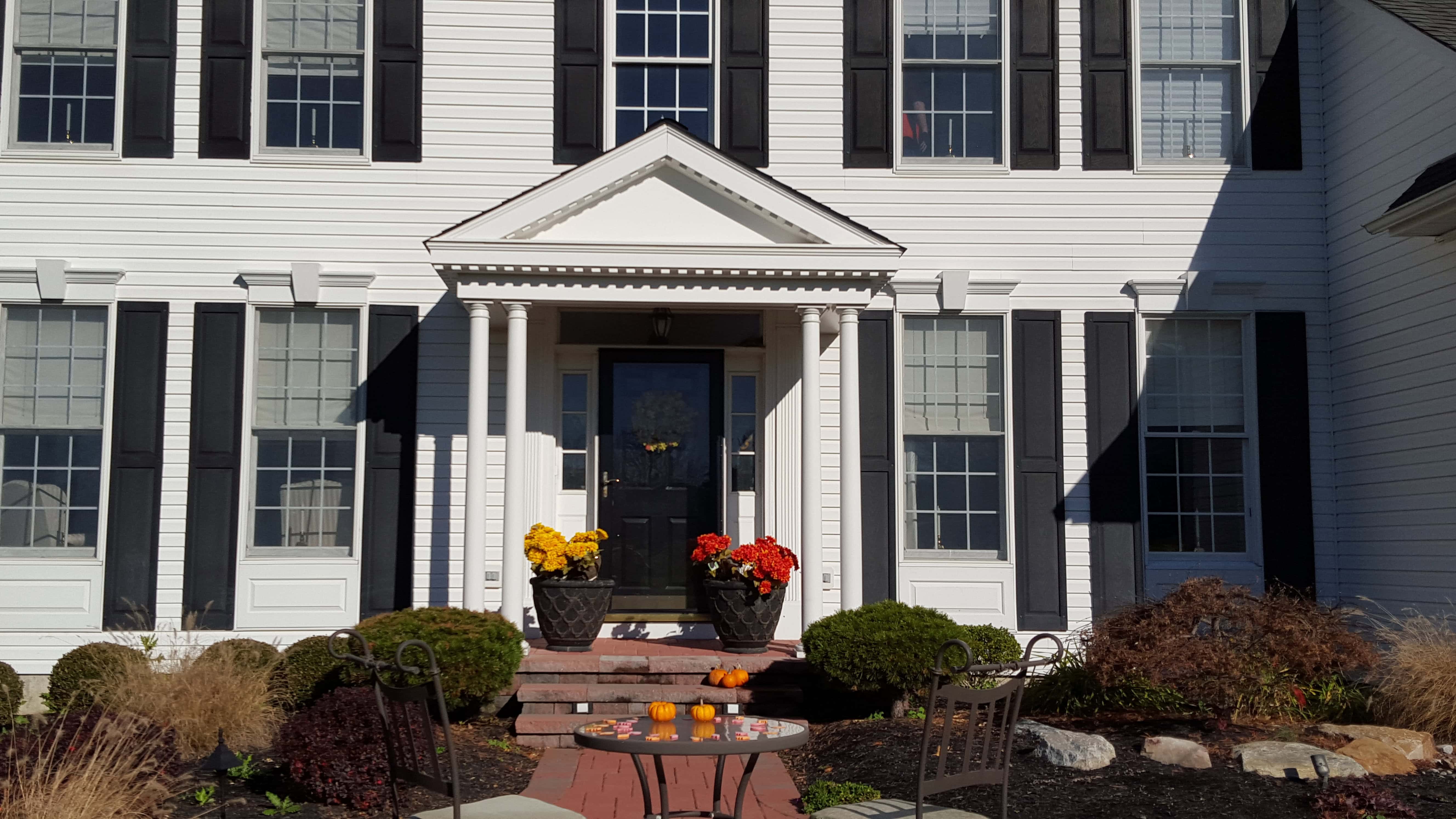A white house with black shutters features a front porch with columns, potted yellow and red flowers on either side of the door, and a small patio table with chairs and orange drinks on a brick walkway.