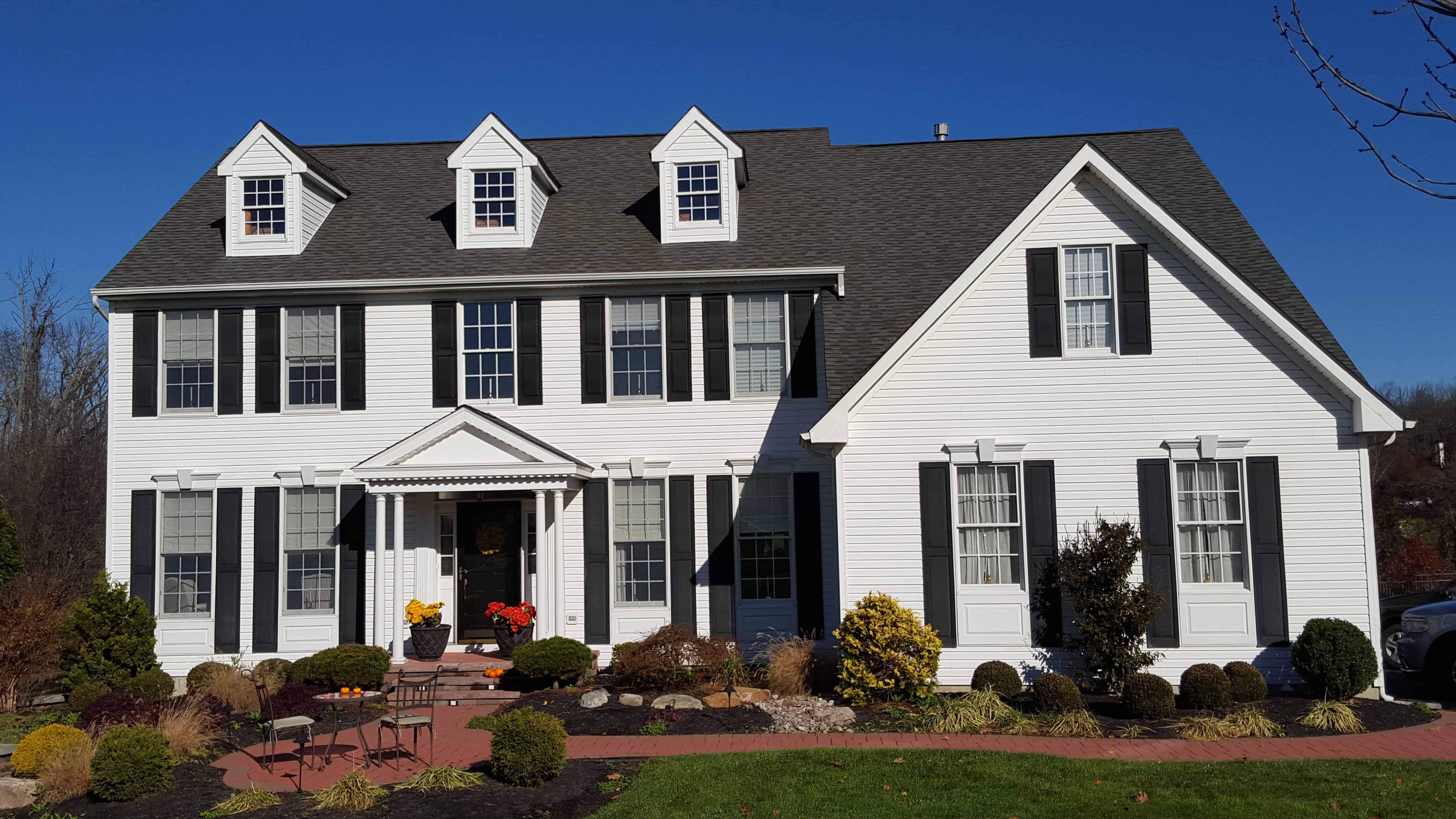 A two-story white house with black shutters, three dormer windows, a dark grey roof, front porch with columns, and neatly landscaped yard with shrubs and a brick path on a sunny day.
