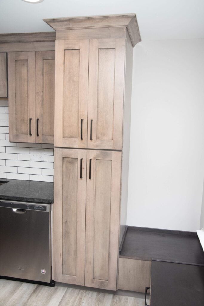 Tall light wood kitchen pantry cabinet with four black handles, set next to a dishwasher and under a crown molding. Dark countertop and white subway tile backsplash are partially visible. Light wood floor below.