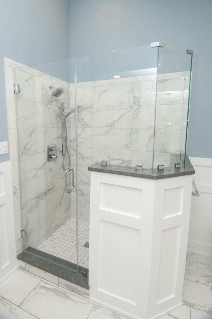 A modern corner shower with glass doors, marble tile walls, a built-in showerhead, and white wood paneling around the base; the bathroom features a light blue wall.