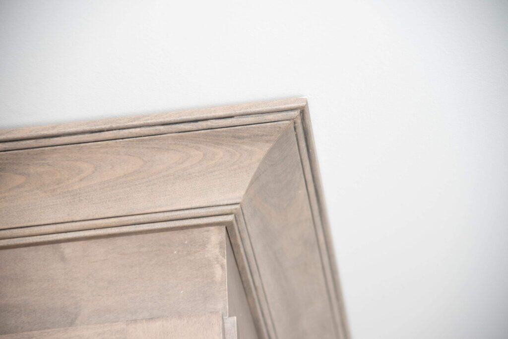Close-up of the top corner of a wooden cabinet with a light finish, showing detailed crown molding against a white wall.