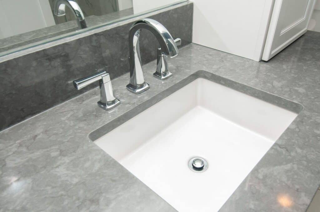 A modern bathroom sink with a rectangular white basin, chrome faucet, and handles set in a gray marble countertop, with a large mirror above and a white cabinet to the side.