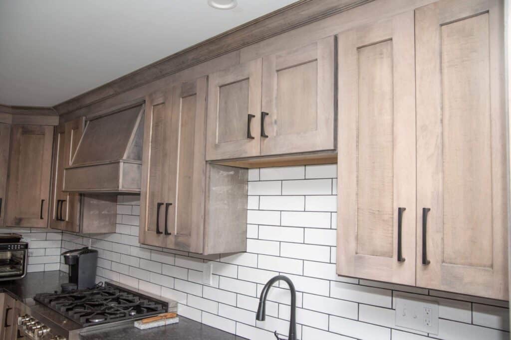 Light wood kitchen cabinets with black handles are mounted above a white subway tile backsplash. A stainless steel sink and gas stove with a dark countertop are visible below the cabinets.