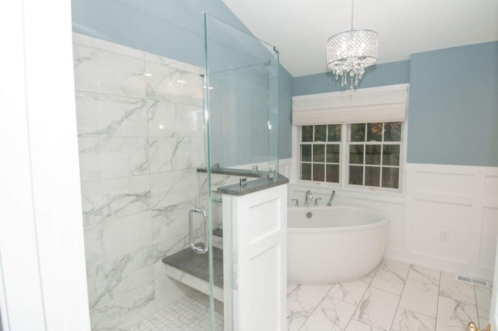Modern bathroom with glass shower, marble tile, freestanding white tub, large window with white blinds, chandelier, and blue walls with white wainscoting.