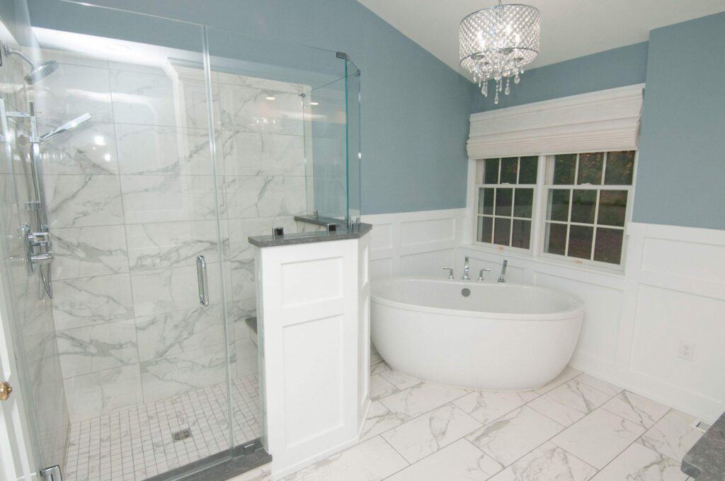 Modern bathroom with a glass-enclosed marble shower, a freestanding oval bathtub, white paneled walls, large window with white blinds, and a decorative chandelier.