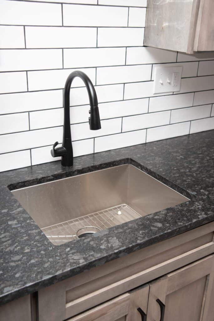 A modern kitchen sink with a black faucet set in a dark granite countertop, light wood cabinets below, and a white subway tile backsplash. An electrical outlet is visible on the wall.