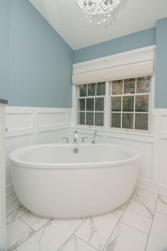 A modern bathroom with a white oval bathtub, marble-tiled floor, light blue walls with white wainscoting, a large window with a white shade, and a crystal chandelier overhead.