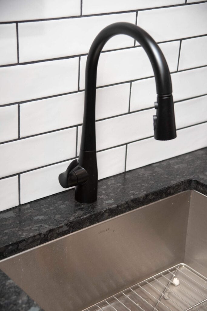 A modern black kitchen faucet over a stainless steel sink, set against a white subway tile backsplash with dark grout. A metal rack is inside the sink.