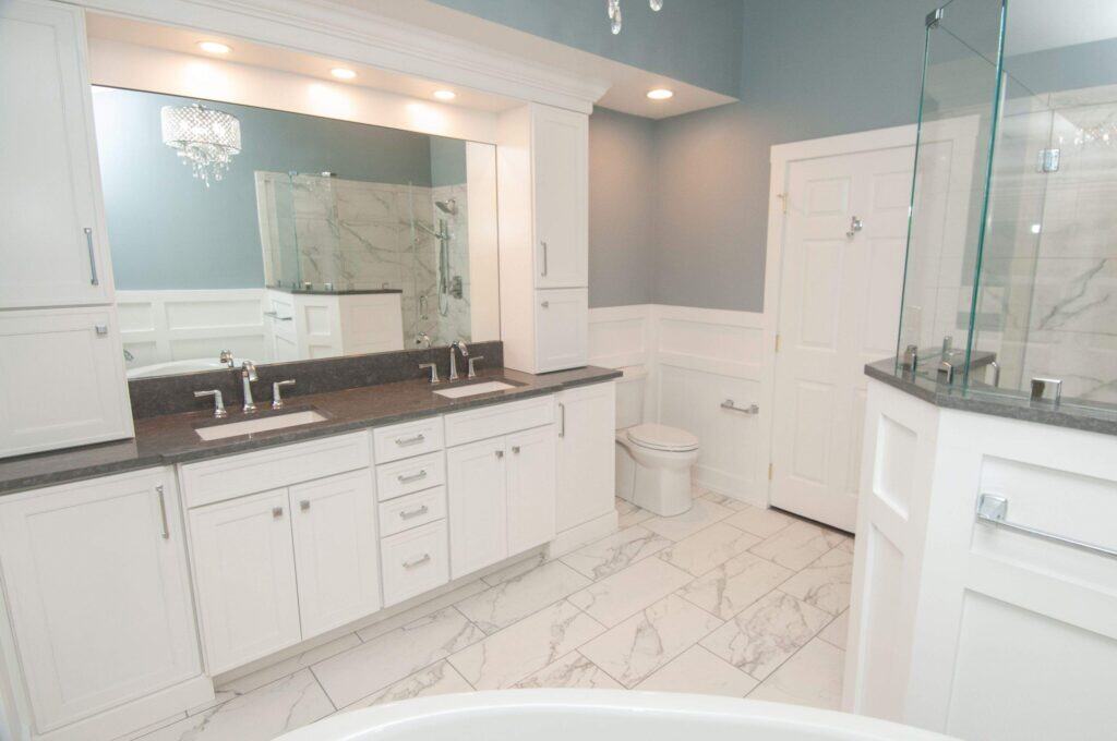 Modern bathroom with white cabinets, double sinks, large mirror, marble tile floor, glass shower, toilet, and soft grey-blue walls. Recessed and chandelier lighting add brightness to the clean, spacious design.